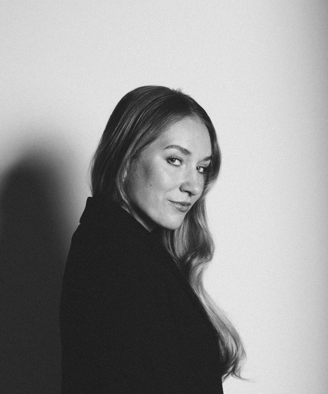 A woman in a violet jacket holds a newspaper, obscuring her face against a light background.